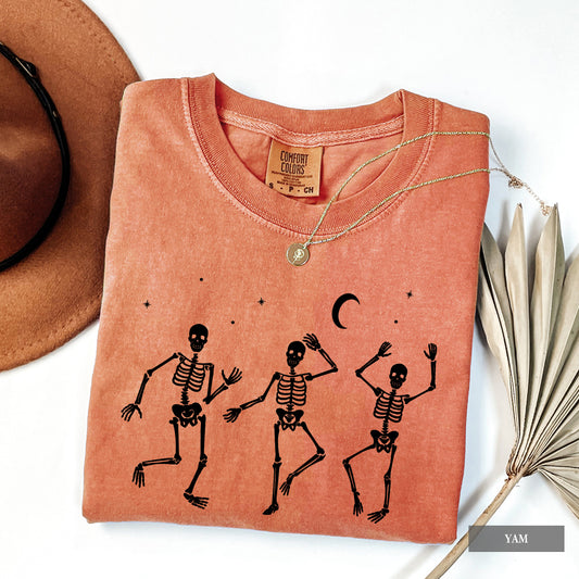 A folded orange comfort colors t-shirt with a black graphic of three dancing skeletons, displayed on a white background with a brown hat, a necklace, and dried palm leaves.