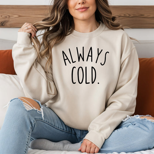 A woman sitting on a bed wearing a cream-colored crewneck sweatshirt with the text 'ALWAYS COLD.' printed in black across the chest, paired with ripped blue jeans.