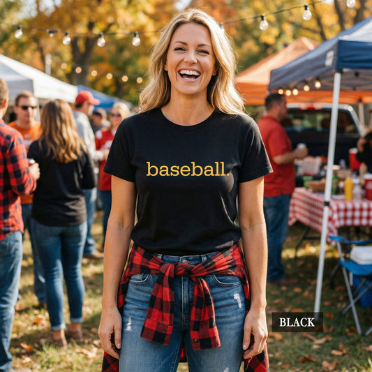 A woman outdoors at a casual autumn gathering wearing a black t-shirt with the word 'baseball' printed in yellow across the chest, blue jeans, and a red plaid flannel shirt tied around her waist. The background shows people, tents, and string lights.