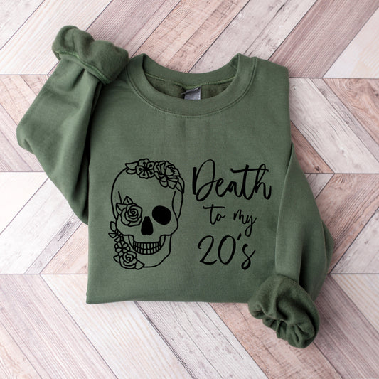 An olive green long-sleeve sweatshirt with a black graphic of a floral sugar skull and the text 'Death to my 20's'. The sweatshirt is laid flat on a light-colored wood plank background.
