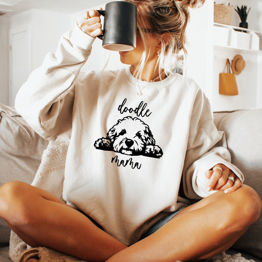 A woman sitting on a couch wearing a cream sweatshirt with a black graphic of a doodle dog and the text 'doodle mama'. She is holding a black mug up to her face in a cozy indoor setting.