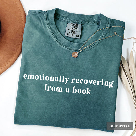 A folded slate blue t-shirt with the text 'emotionally recovering from a book' in white lowercase letters, styled with a necklace and props on a white background.