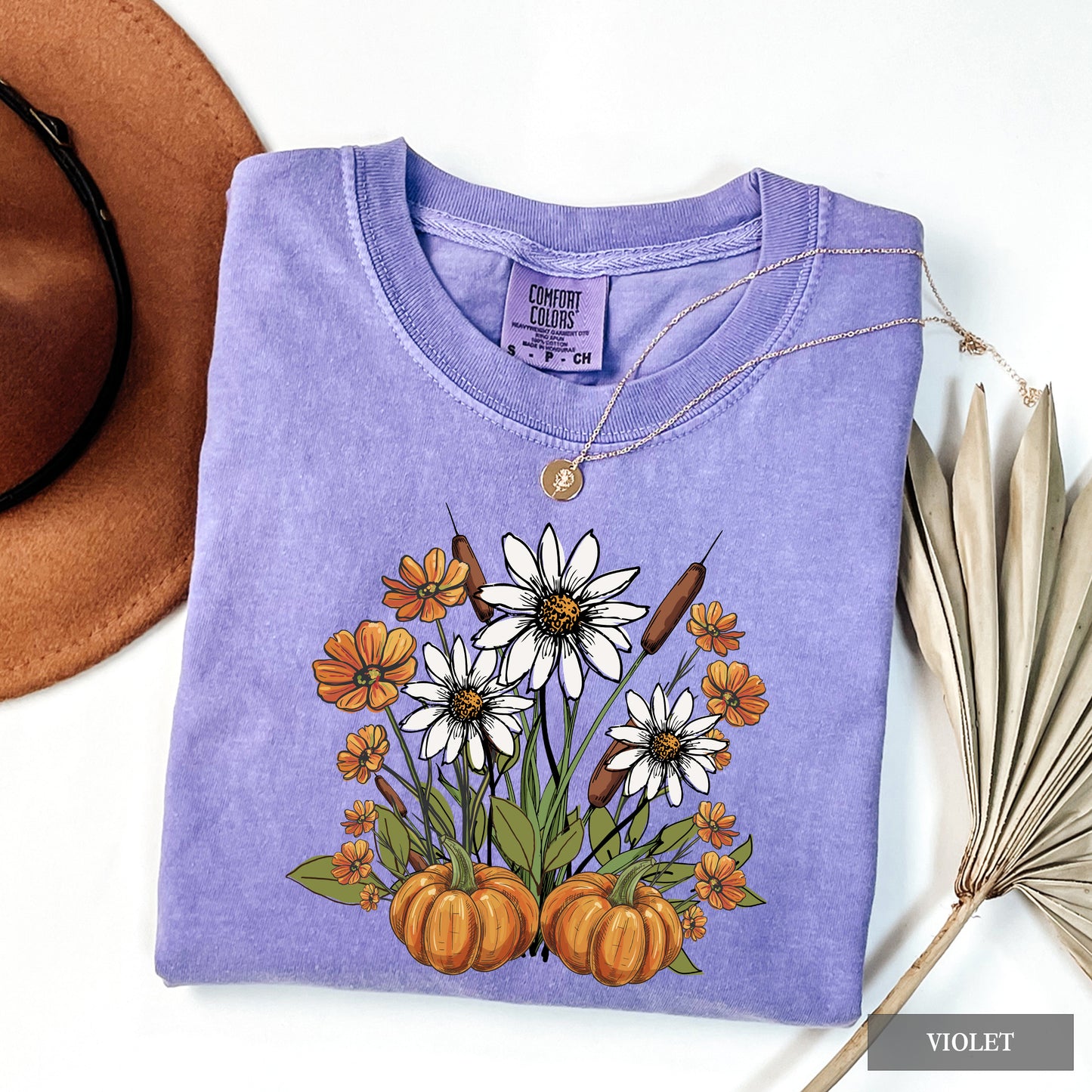 Folded purple shirt with floral and pumpkin design, next to a brown hat and dried leaves.