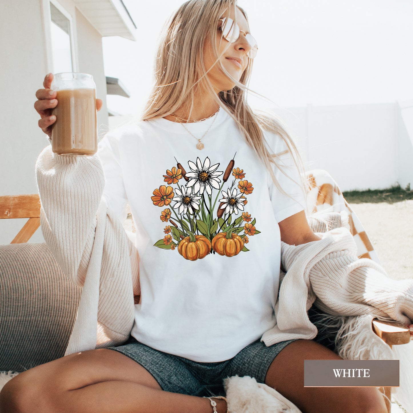 Woman wearing a white t-shirt with pumpkins and flowers design, holding a drink outdoors.