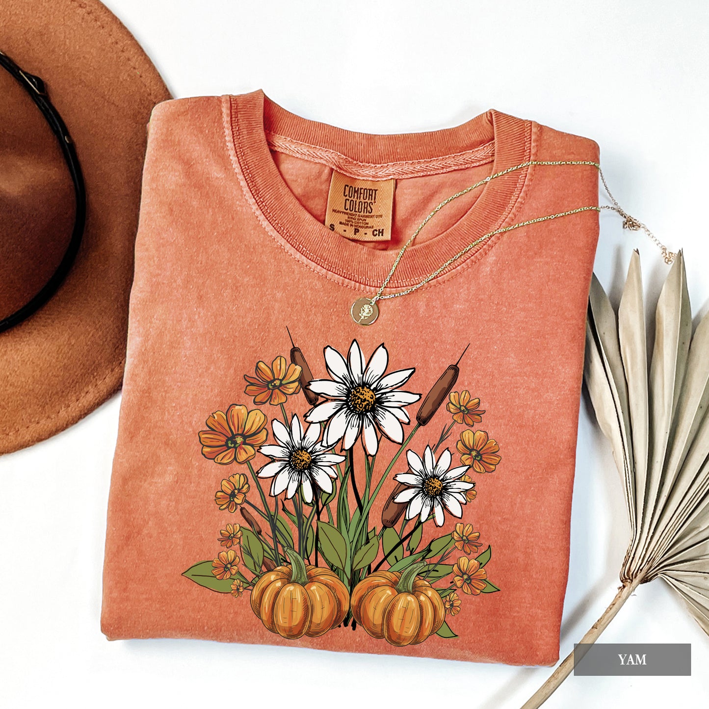 Folded orange t-shirt with floral and pumpkin design, next to a brown hat and dried leaves.