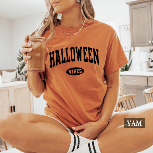 A woman wearing a burnt orange graphic t-shirt with 'HALLOWEEN VIBES' printed in black on the front, holding an iced coffee in a bright kitchen setting. A YAM brand label is visible in the bottom right corner.
