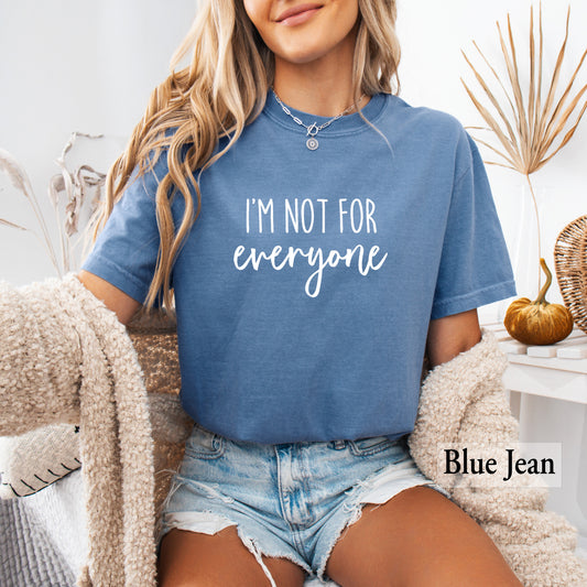 A woman wearing a heathered blue crewneck t-shirt with the words 'I'm Not For Everyone' printed in white across the chest, paired with denim shorts and a fuzzy cream cardigan, seated in a styled indoor setting.
