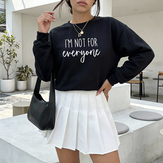 A woman wearing a black crewneck sweatshirt with the white text 'I'M NOT FOR everyone' and a white pleated mini skirt, carrying a black shoulder bag in an outdoor patio setting.