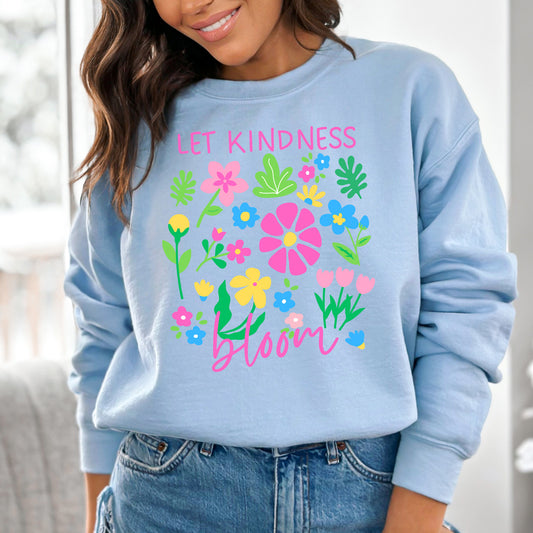 Light blue sweatshirt with colorful floral design and text 'Let kindness bloom' worn by a person.