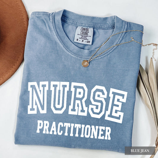 A folded light blue t-shirt with 'NURSE PRACTITIONER' printed in white varsity letters, styled with a gold necklace and a brown hat on a white background.