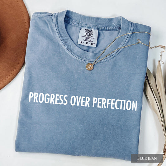 A folded light blue t-shirt with the phrase 'PROGRESS OVER PERFECTION' printed in white across the chest. The shirt is styled in a flat lay with a gold necklace, a brown hat edge, and dried leaves on a white background.