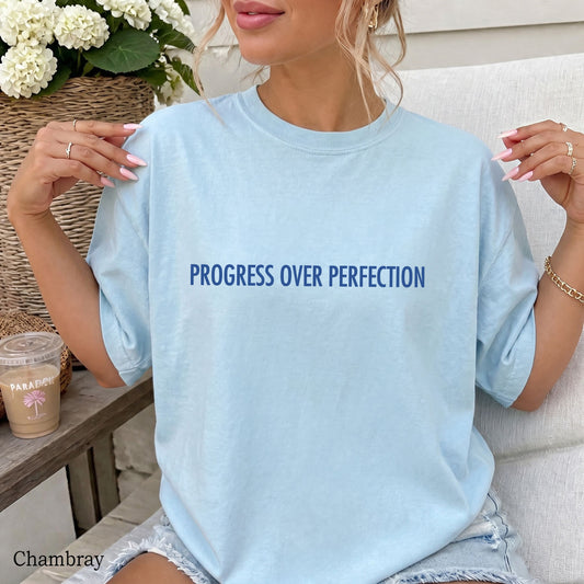 A woman wearing a light chambray blue oversized t-shirt with the text 'PROGRESS OVER PERFECTION' printed in dark blue across the chest. She is sitting on a white chair next to a table with a coffee cup and a basket of white flowers.