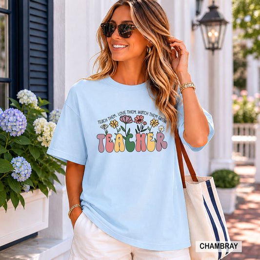 Woman wearing a light blue t-shirt with 'Teacher' and floral design, standing outdoors.