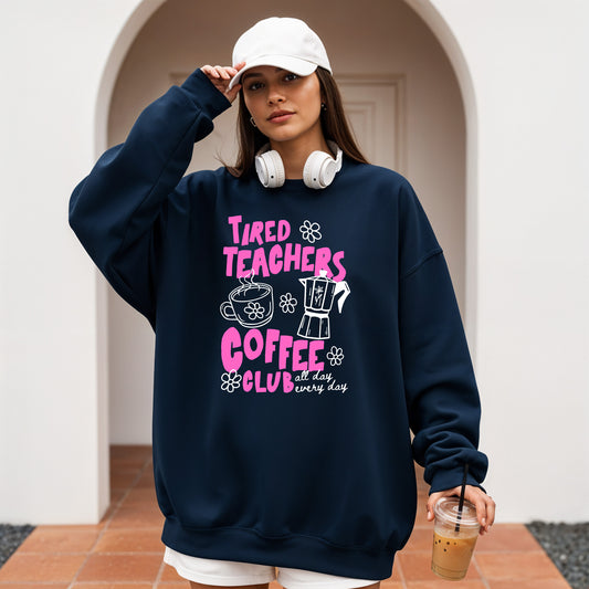 Person wearing a navy sweatshirt with 'Tired Teachers Coffee Club' text, holding a drink.