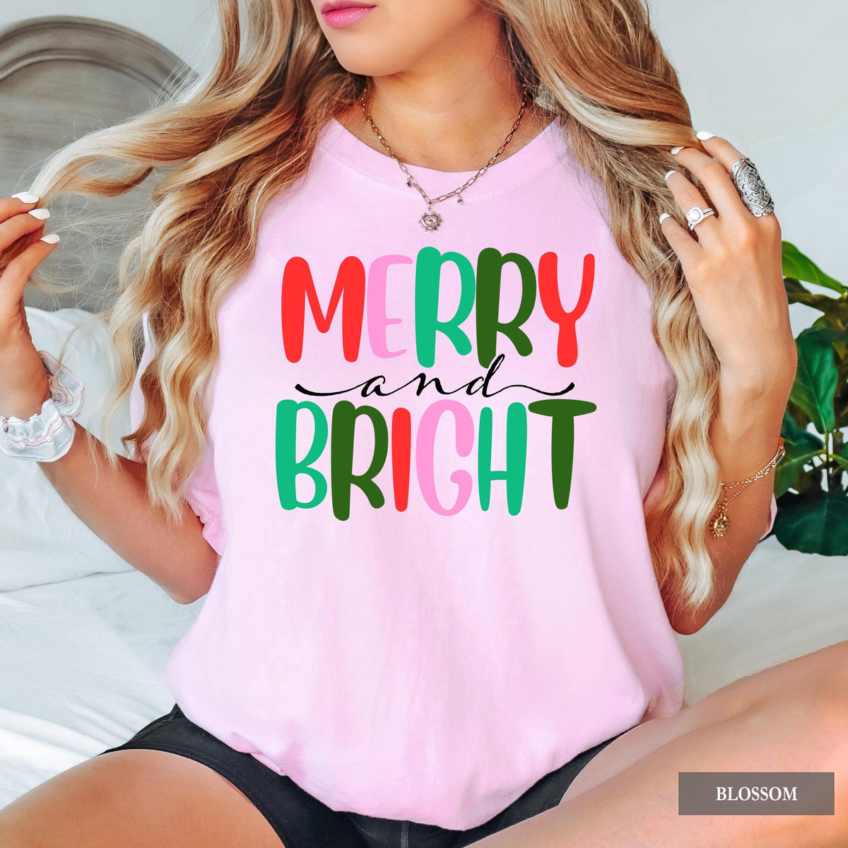 A person wearing a light pink t-shirt with colorful Merry and Bright text against a white background.