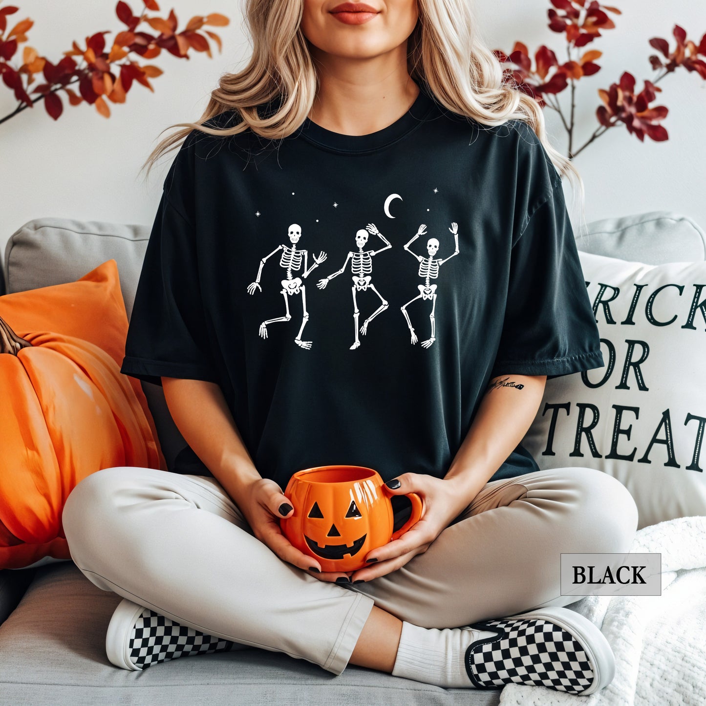 A woman sitting on a couch wearing a black graphic t-shirt with three white dancing skeletons under a crescent moon, holding an orange jack-o'-lantern mug. The background features a pumpkin, pillows, and autumn leaves.