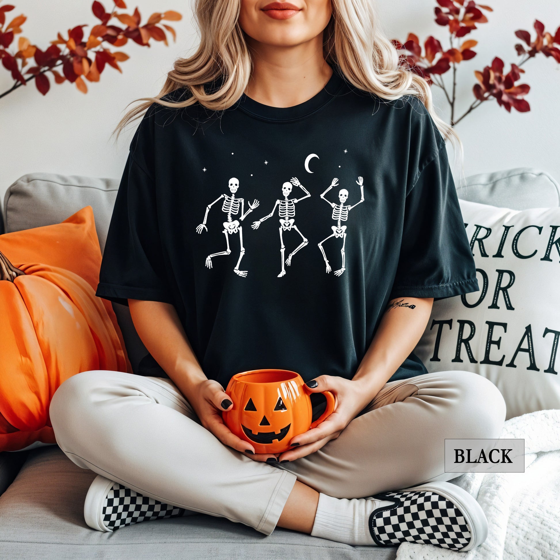 A woman sitting on a couch wearing a black graphic t-shirt with three white dancing skeletons under a crescent moon, holding an orange jack-o'-lantern mug. The background features a pumpkin, pillows, and autumn leaves.