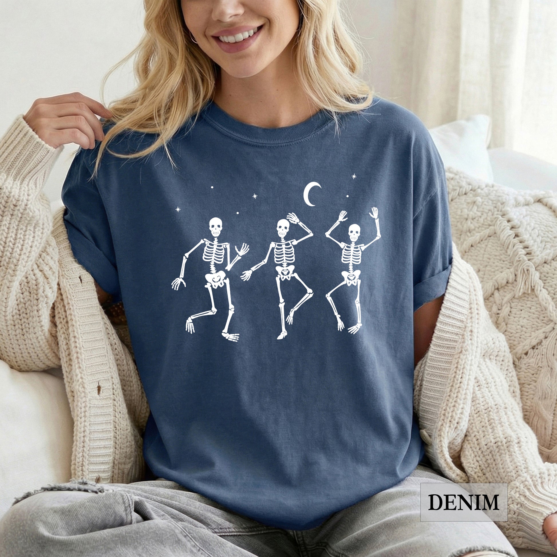 A woman wearing a navy blue t-shirt with a white graphic of three dancing skeletons under a crescent moon, sitting on a couch in a cozy setting.