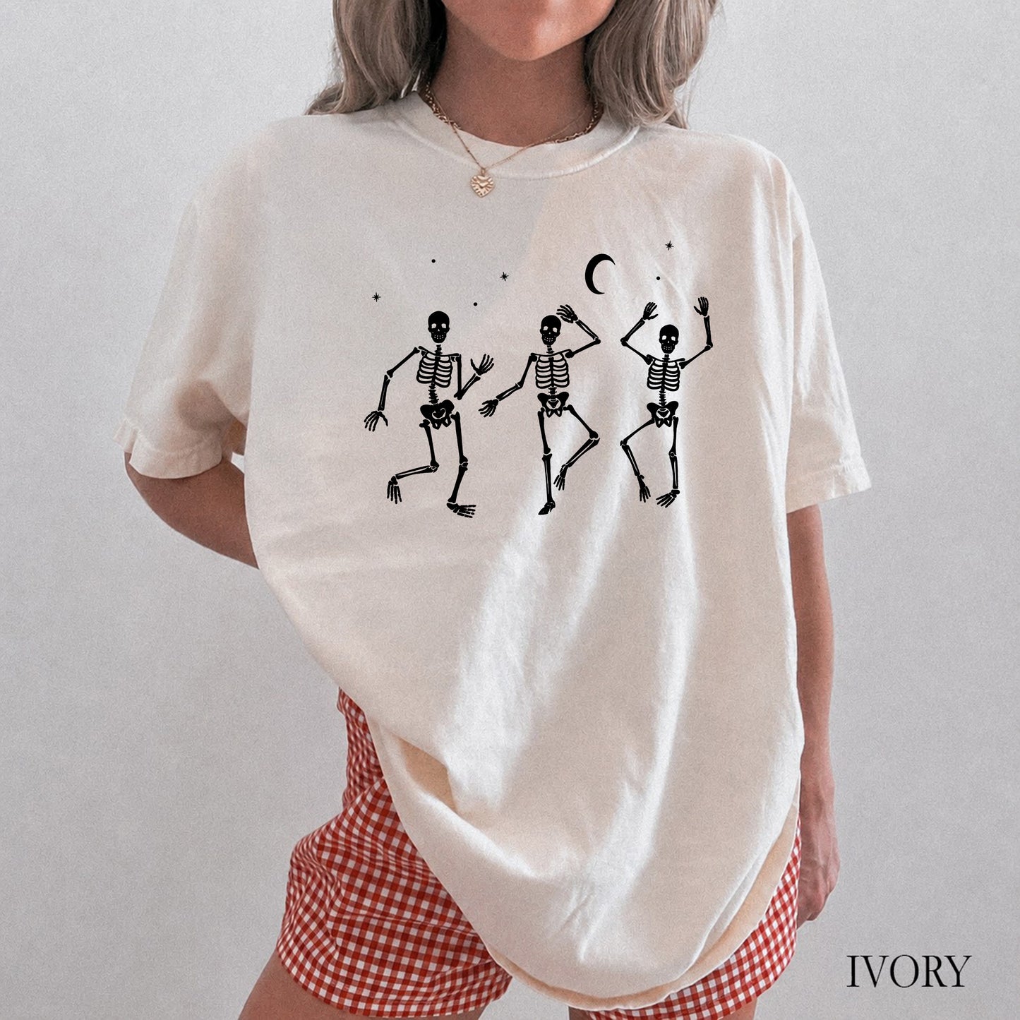 A woman wearing an oversized ivory short-sleeve t-shirt with a black graphic of three dancing skeletons under a crescent moon, paired with red and white gingham shorts. The word 'IVORY' is printed in the bottom right corner.