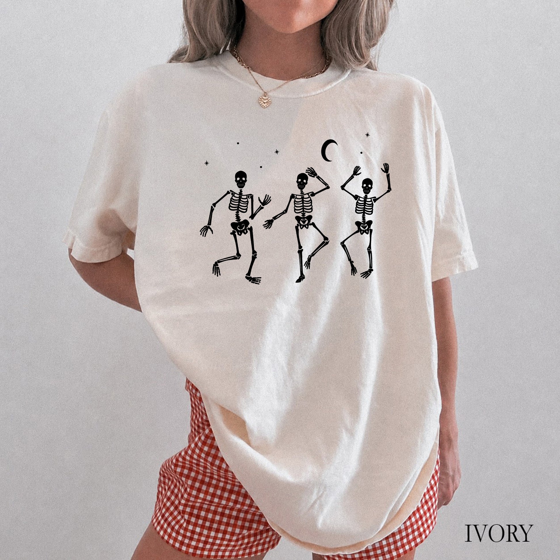 A woman wearing an oversized ivory short-sleeve t-shirt with a black graphic of three dancing skeletons under a crescent moon, paired with red and white gingham shorts. The word 'IVORY' is printed in the bottom right corner.