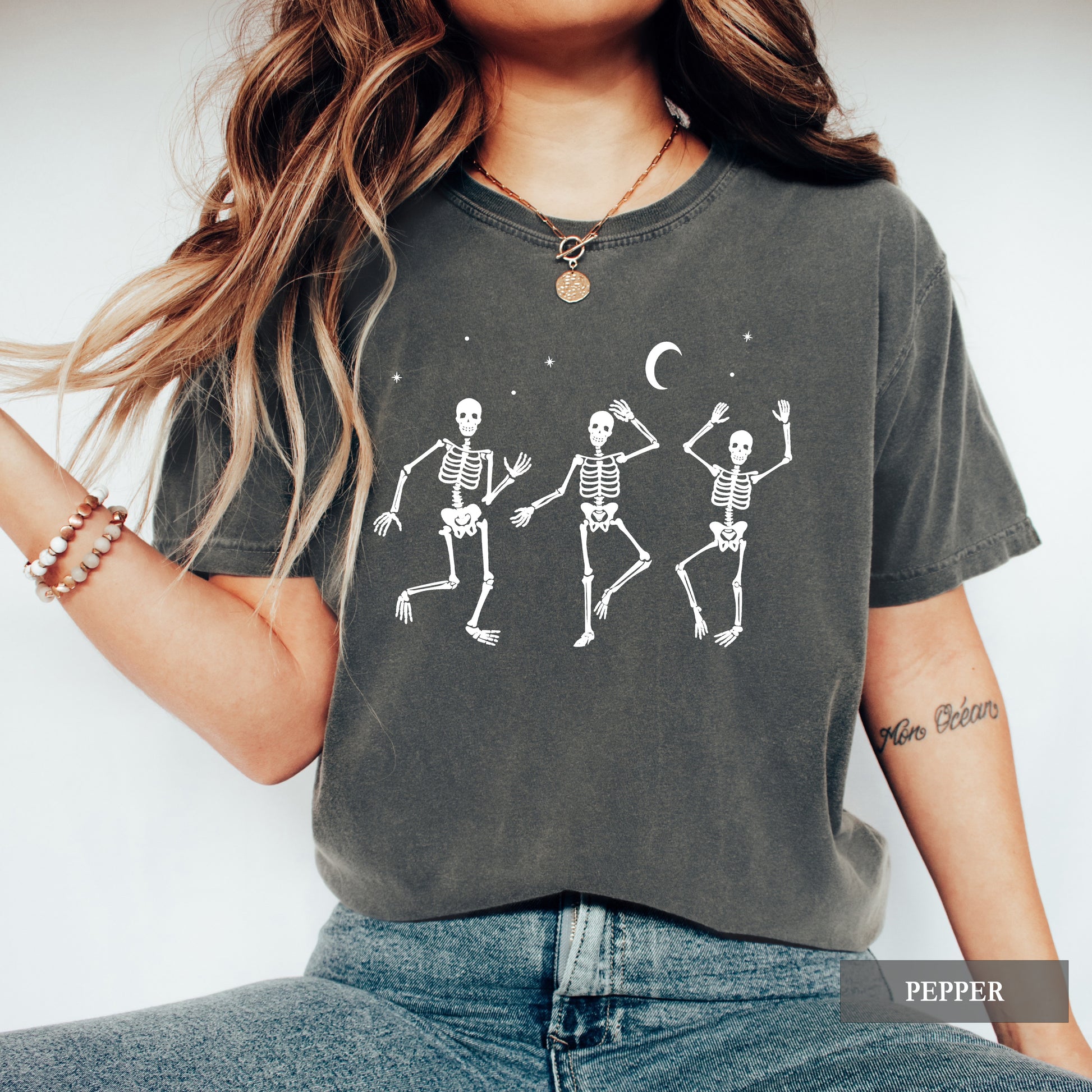 A woman wearing a dark charcoal gray t-shirt with a white graphic of three dancing skeletons under a crescent moon, paired with blue jeans.