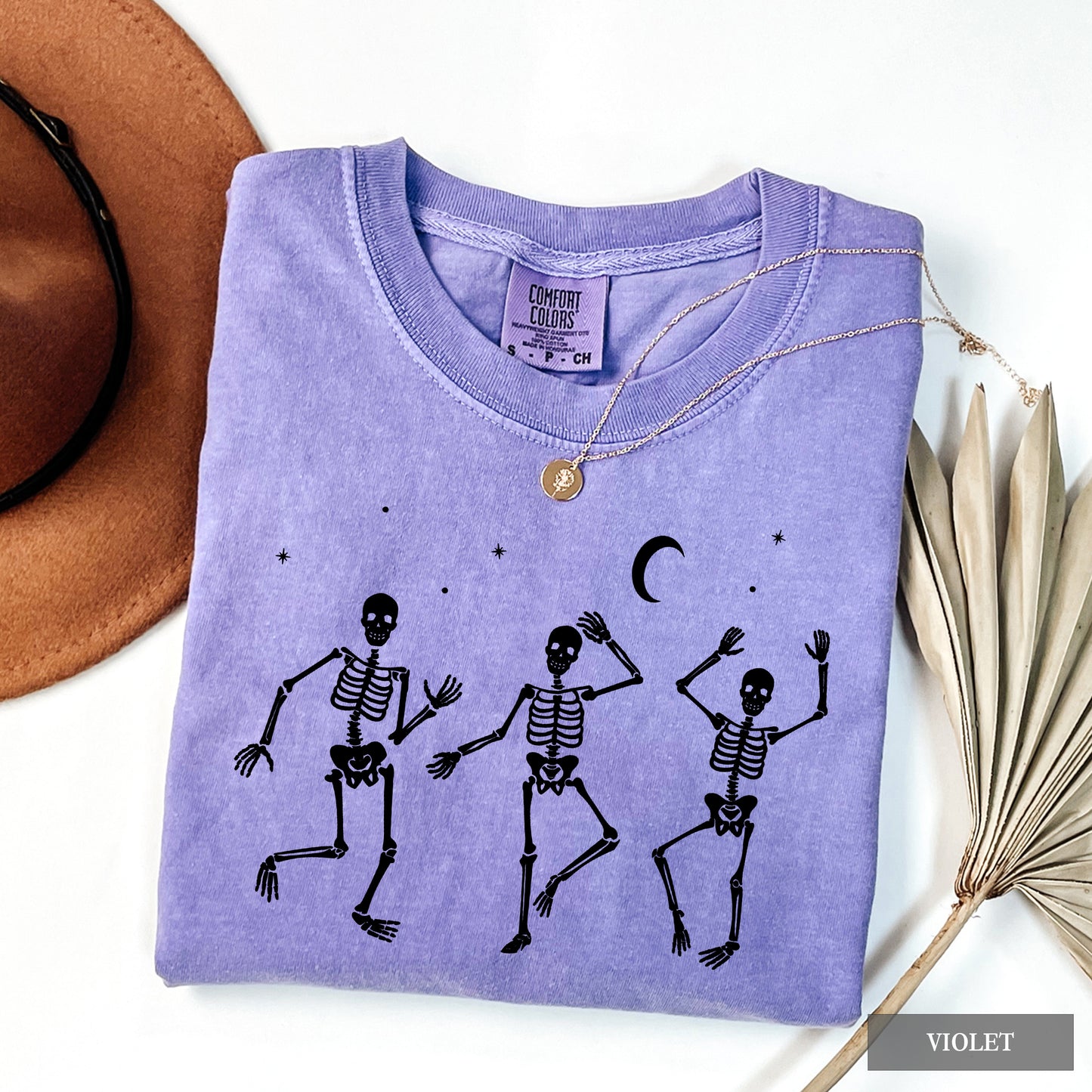 A folded lavender-purple t-shirt with a black graphic of three dancing skeletons under a crescent moon and stars. The shirt is styled in a flat lay with a brown hat, sunglasses, a gold necklace, and dried palm leaves on a white background.