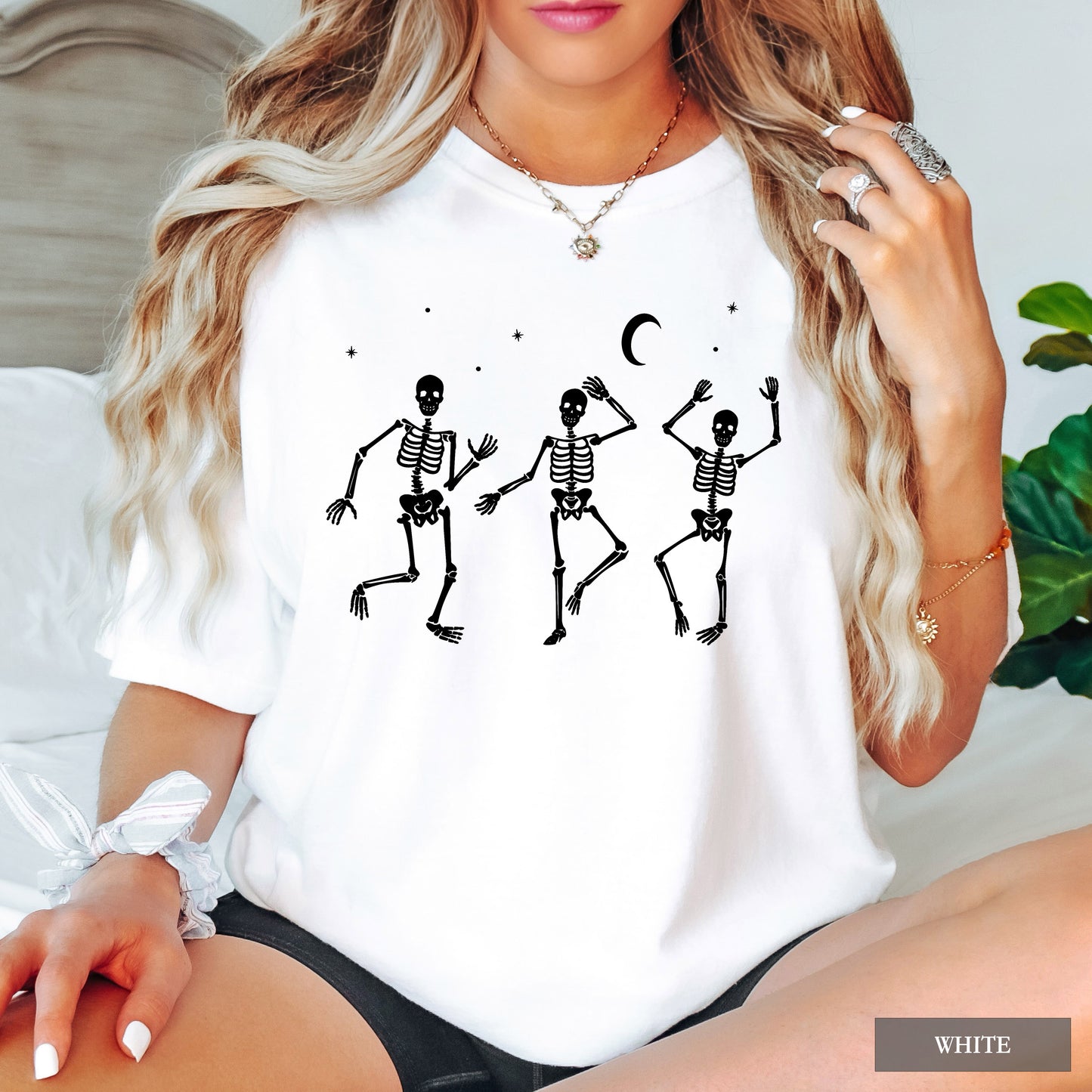 A woman wearing a white short-sleeve t-shirt with a black graphic of three dancing skeletons under a crescent moon and stars. The image is a lifestyle shot with a 'WHITE' label overlay in the bottom right corner.