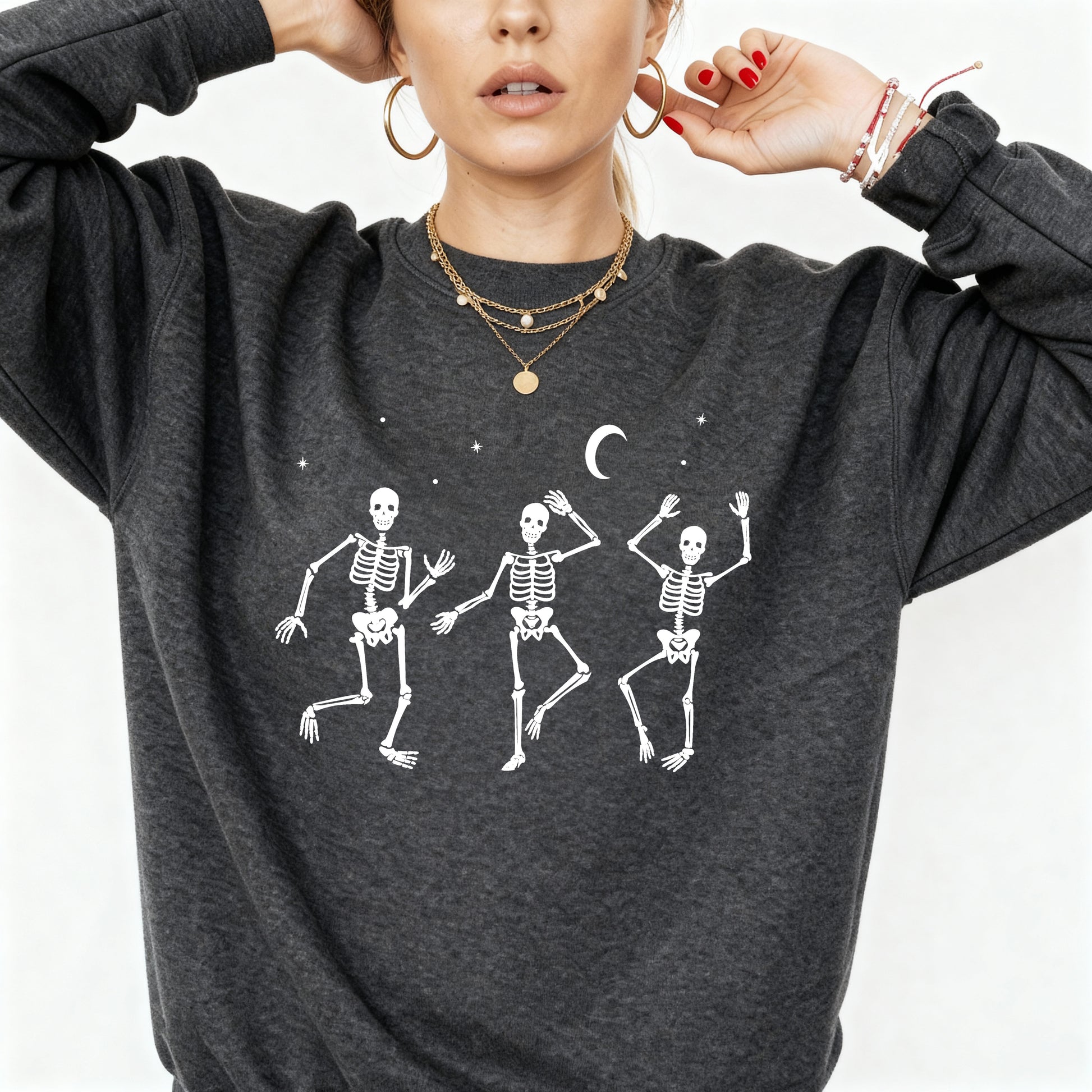 A woman wearing a dark heather gray sweatshirt with a white graphic of three dancing skeletons under a crescent moon and stars.