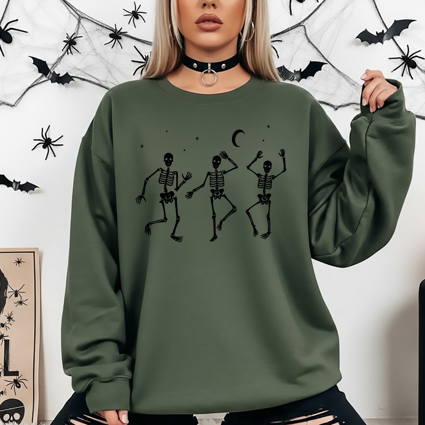 A woman wearing an oversized olive green sweatshirt with a black graphic of three dancing skeletons under a crescent moon, posed against a white wall decorated with black spiderwebs and bats.