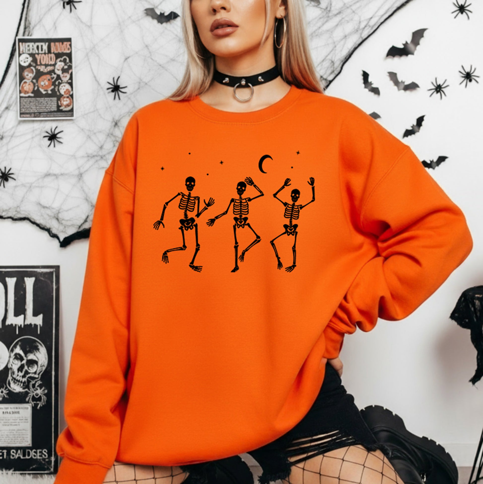 A woman wearing a bright orange crewneck sweatshirt with a black graphic of three dancing skeletons under a crescent moon, posed against a Halloween-themed wall with spiderwebs, bats, and posters.