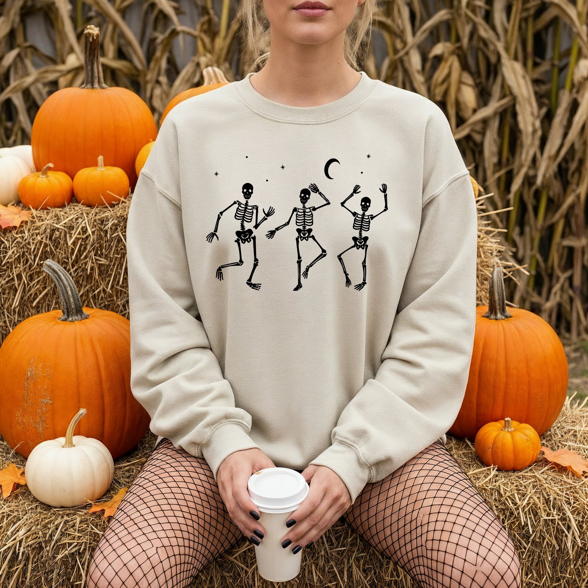 A woman wearing a light beige crewneck sweatshirt with a black graphic of three dancing skeletons under a crescent moon, sitting on a hay bale surrounded by pumpkins.