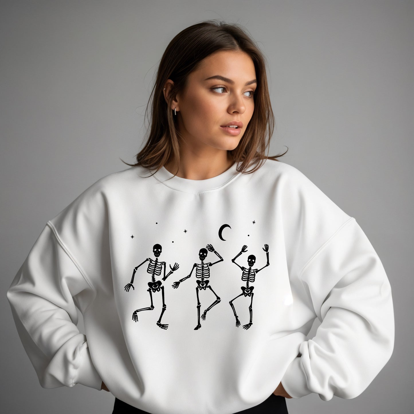 A woman wearing a white crewneck sweatshirt with a black graphic of three dancing skeletons under a crescent moon, photographed against a neutral gray studio background.