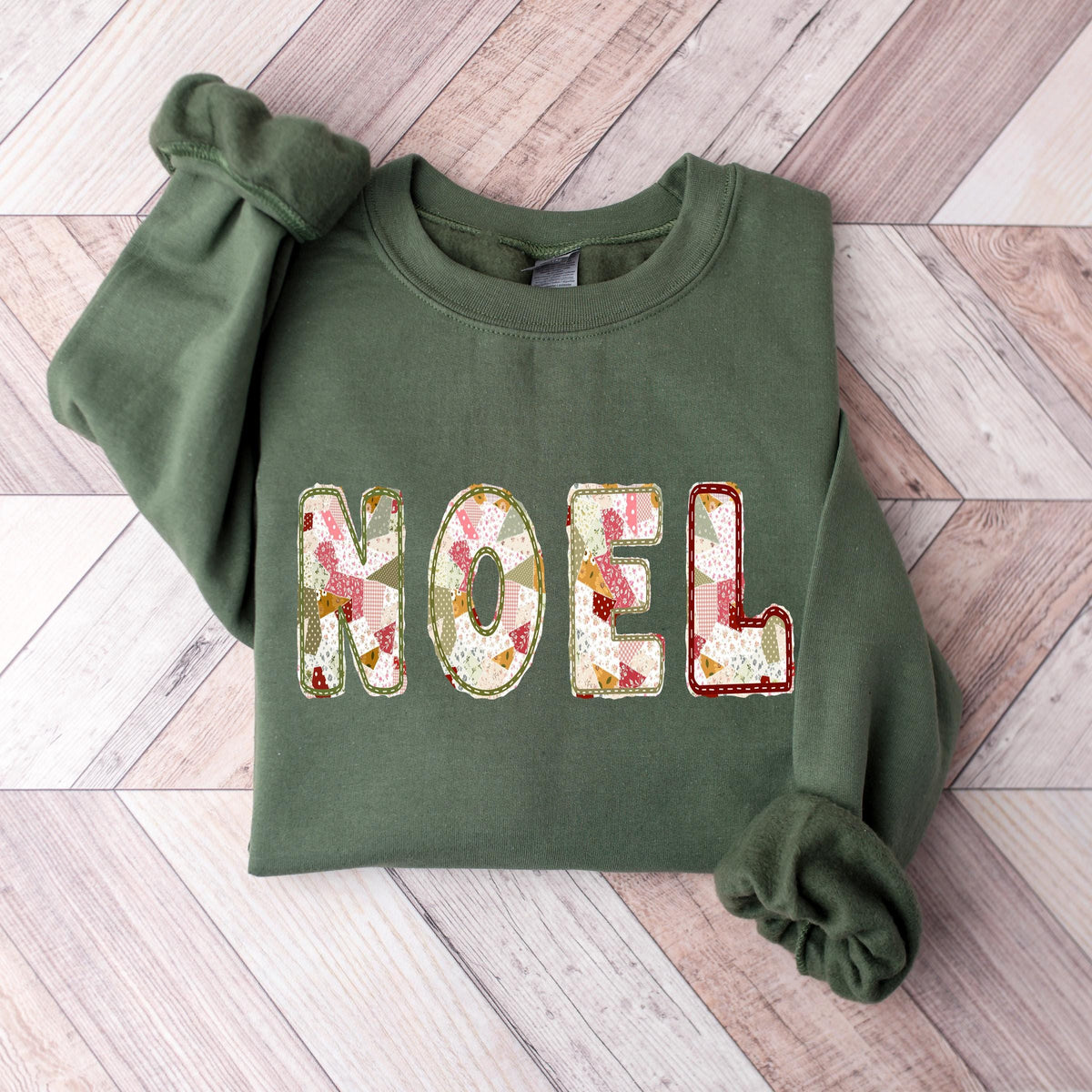 A military green Gildan crewneck sweatshirt with NOEL printed on it in a muted sage, maroon, and white patchwork pattern.