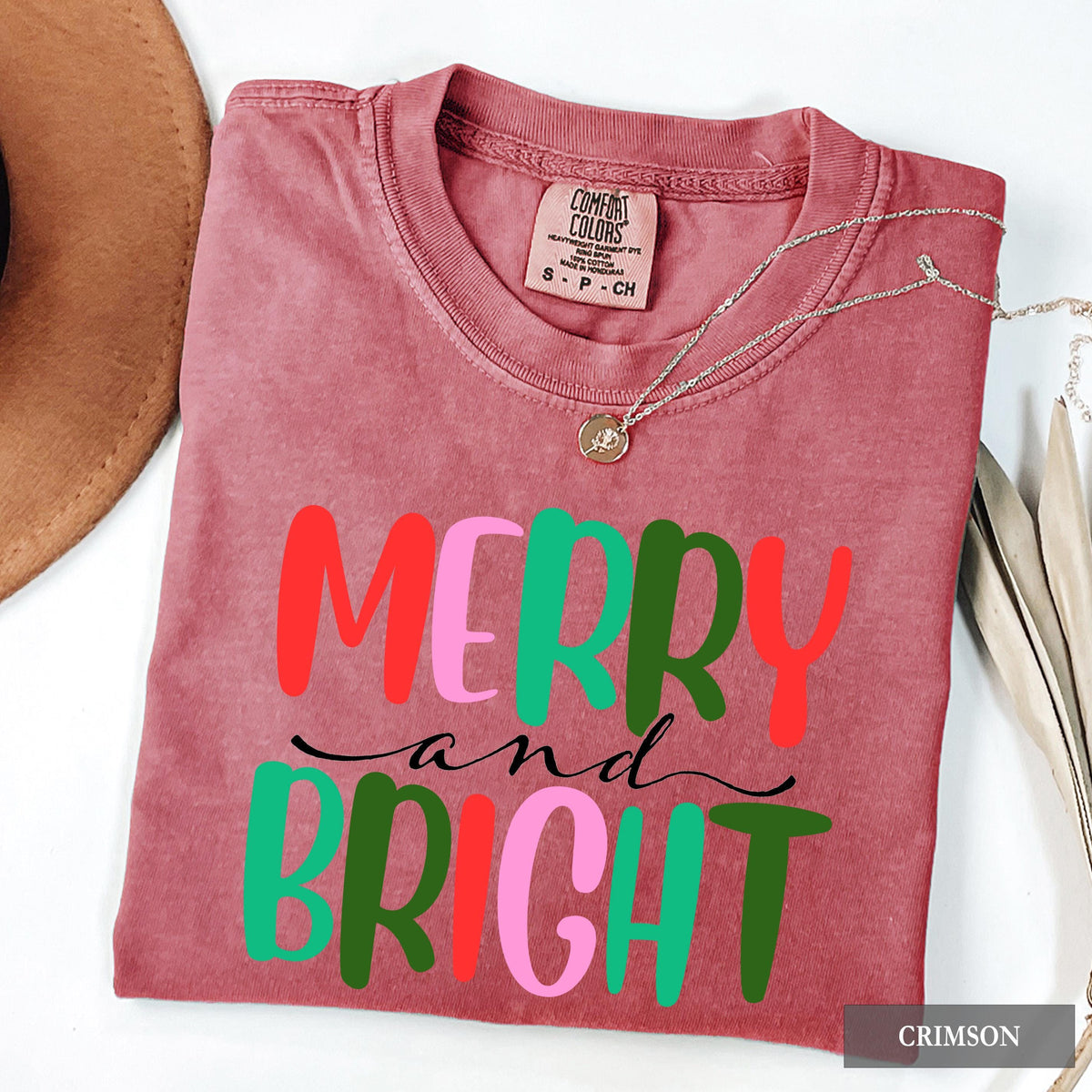 A flat lay of a crimson t-shirt featuring colorful Merry and Bright text, next to a brown hat and plant leaves.