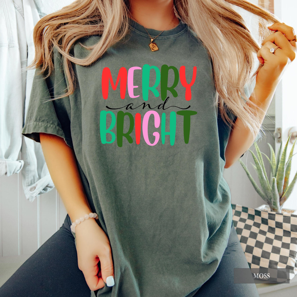 A person wearing a dark grey t-shirt with colorful Merry and Bright text, holding a Santa mug, next to a lit Christmas tree.