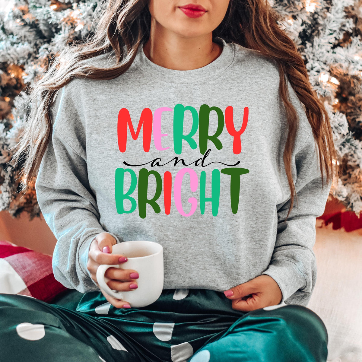A woman holds a white mug while wearing a gray sweatshirt with colorful Merry and Bright lettering.