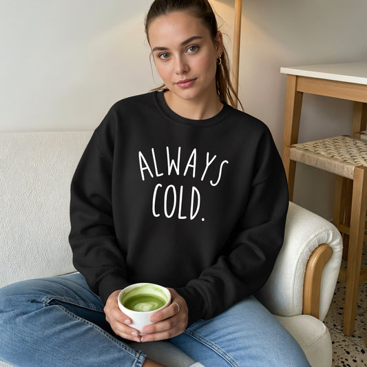 A woman sitting on a sofa wearing a black crewneck sweatshirt with the text 'ALWAYS COLD.' printed in white across the chest, paired with blue jeans.