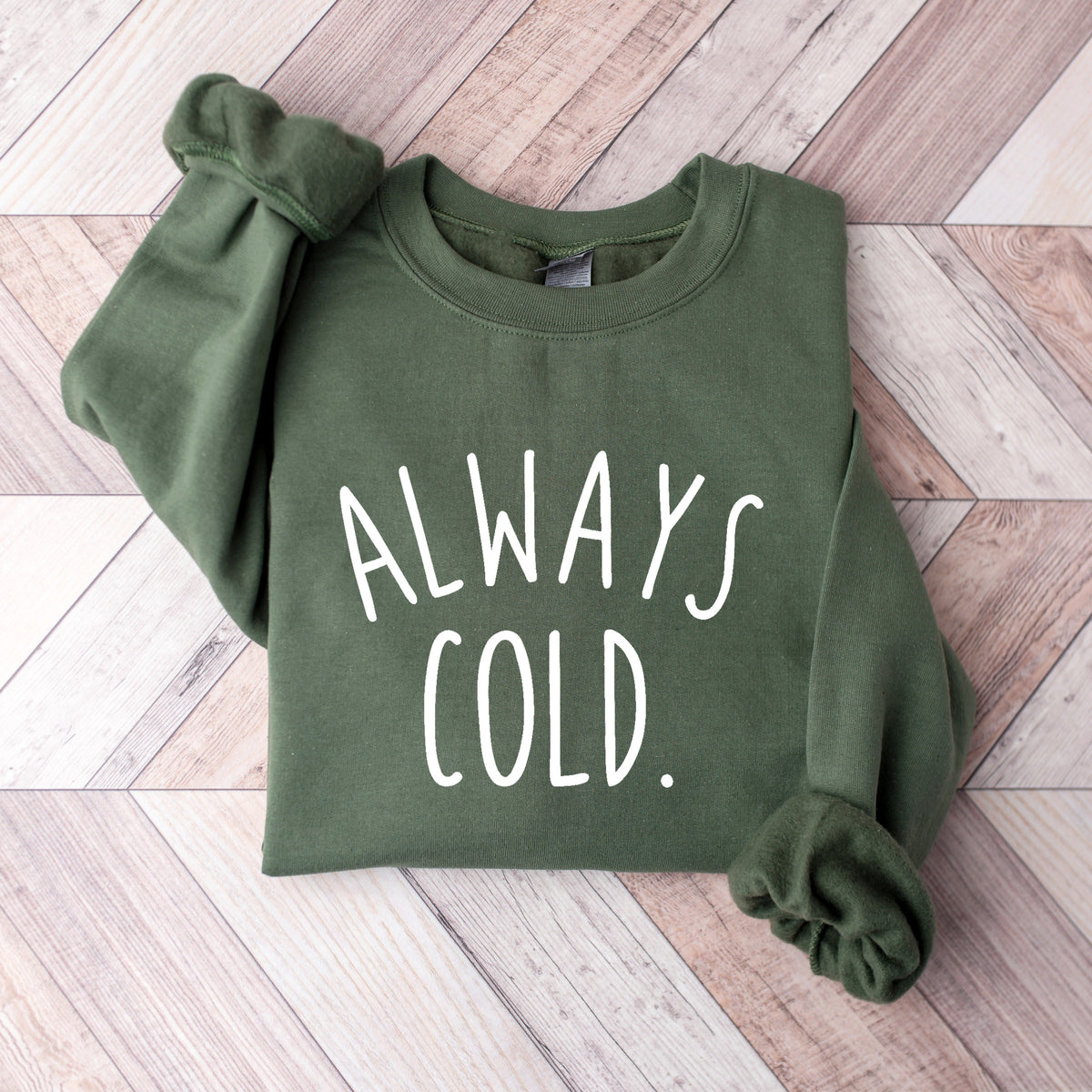 An olive green crewneck sweatshirt with the phrase 'ALWAYS COLD.' printed in white across the chest, laid flat on a light-colored wood plank background.