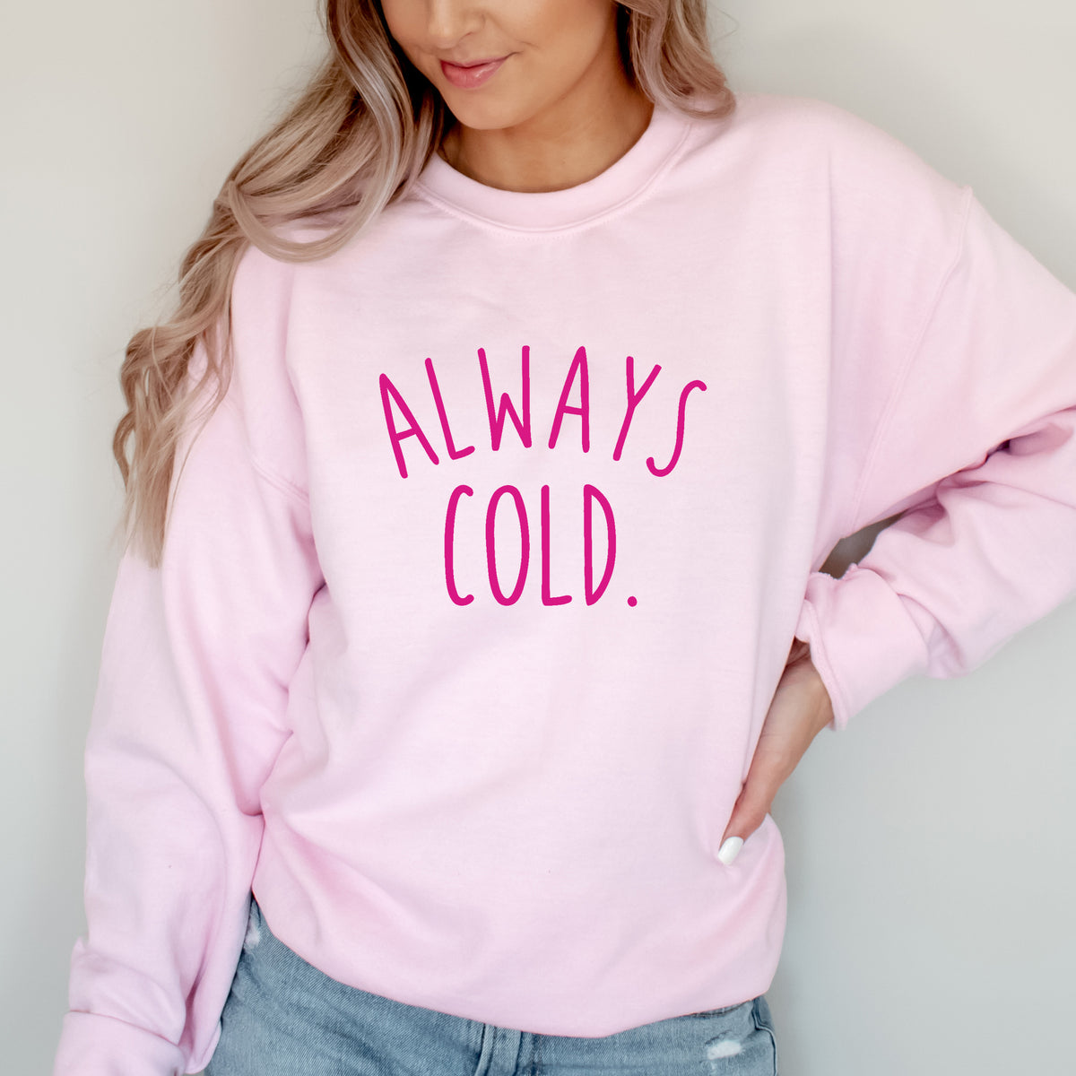 A woman wearing a light pink crewneck sweatshirt with the text 'ALWAYS COLD.' printed in hot pink across the chest, paired with ripped blue jeans.