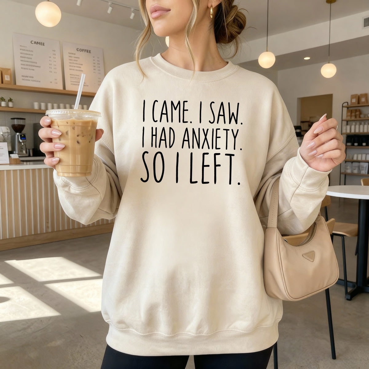 A woman wearing a sand sweatshirt that says 'I Came I Saw I Had Anxiety So I Left.'