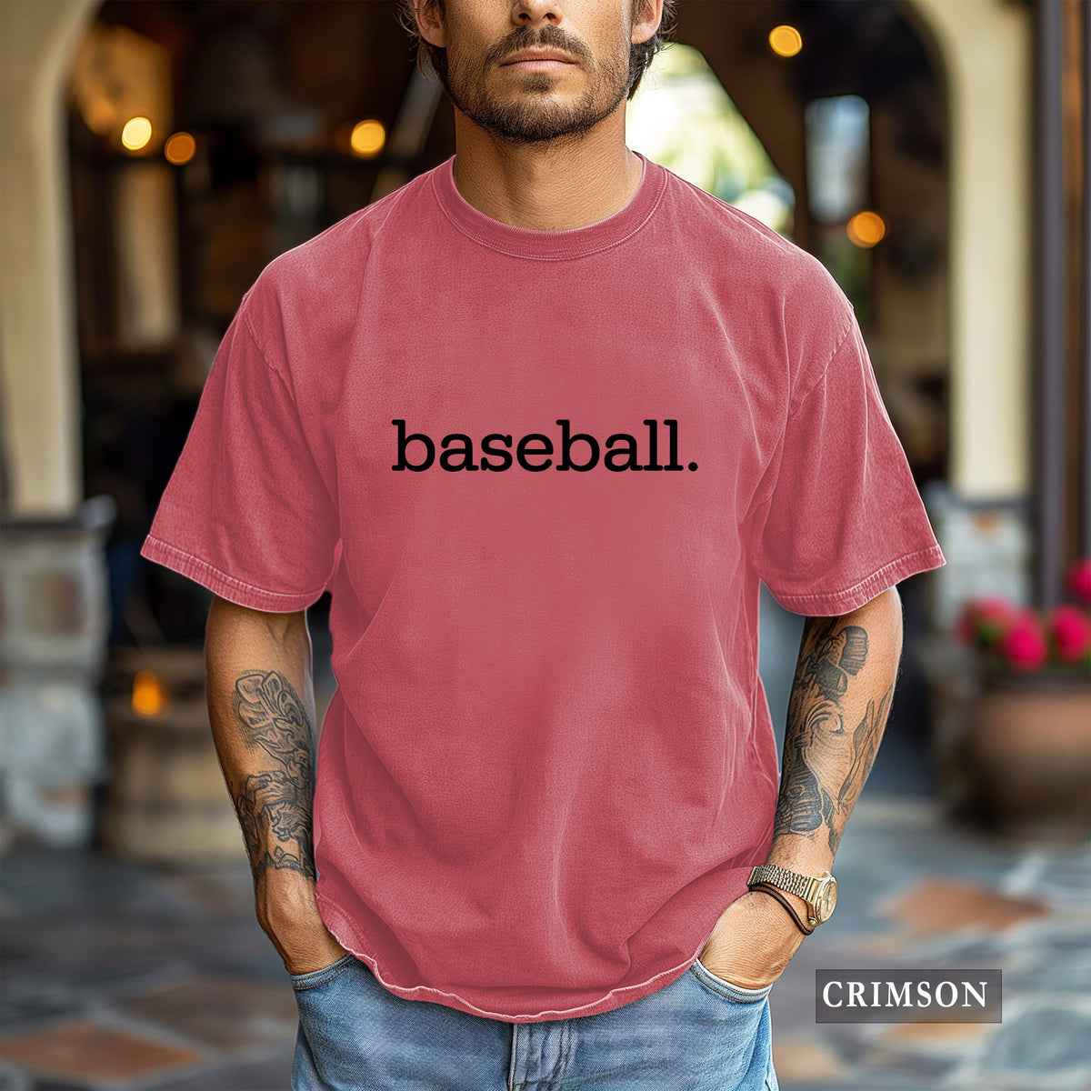 A man wearing a faded crimson crewneck t-shirt with the word 'baseball.' printed in black across the chest, set against a blurred outdoor patio background.