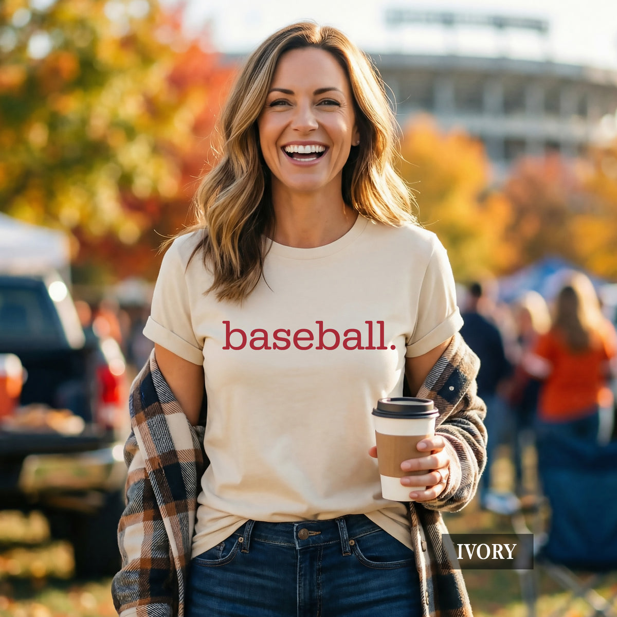 A woman outdoors wearing an ivory short-sleeve T-shirt with the word 'baseball.' printed in red across the chest, holding a coffee cup and wearing a plaid shirt draped off her shoulders.