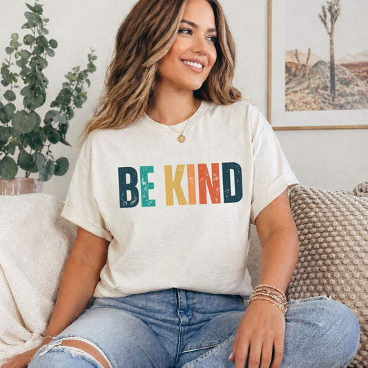 Woman wearing a 'BE KIND' t-shirt sitting on a couch in a cozy living room.