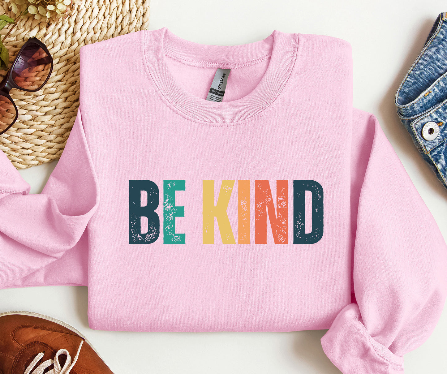 Pink sweatshirt with 'BE KIND' in colorful letters on a white surface.
