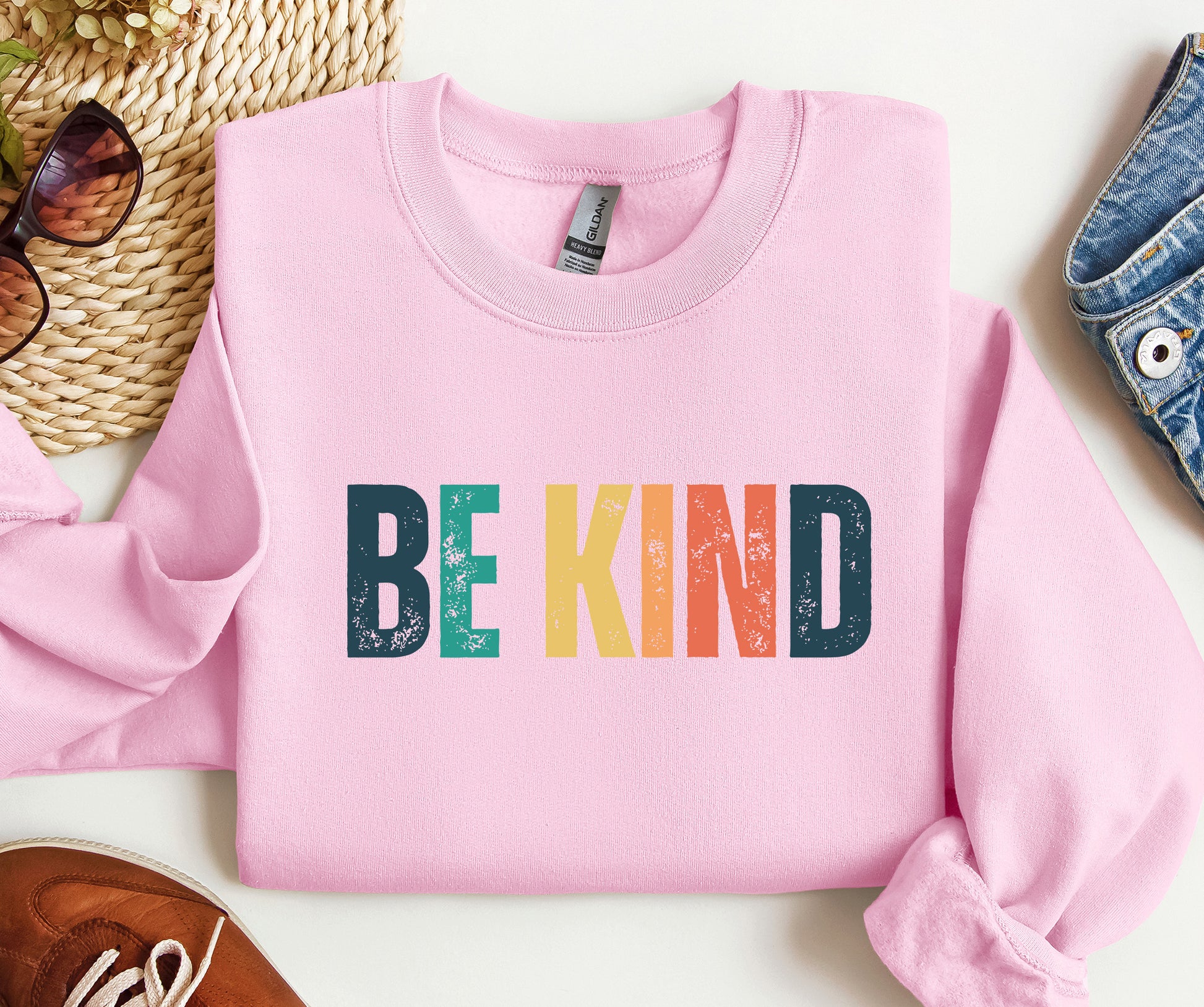 Pink sweatshirt with 'BE KIND' in colorful letters on a white surface.