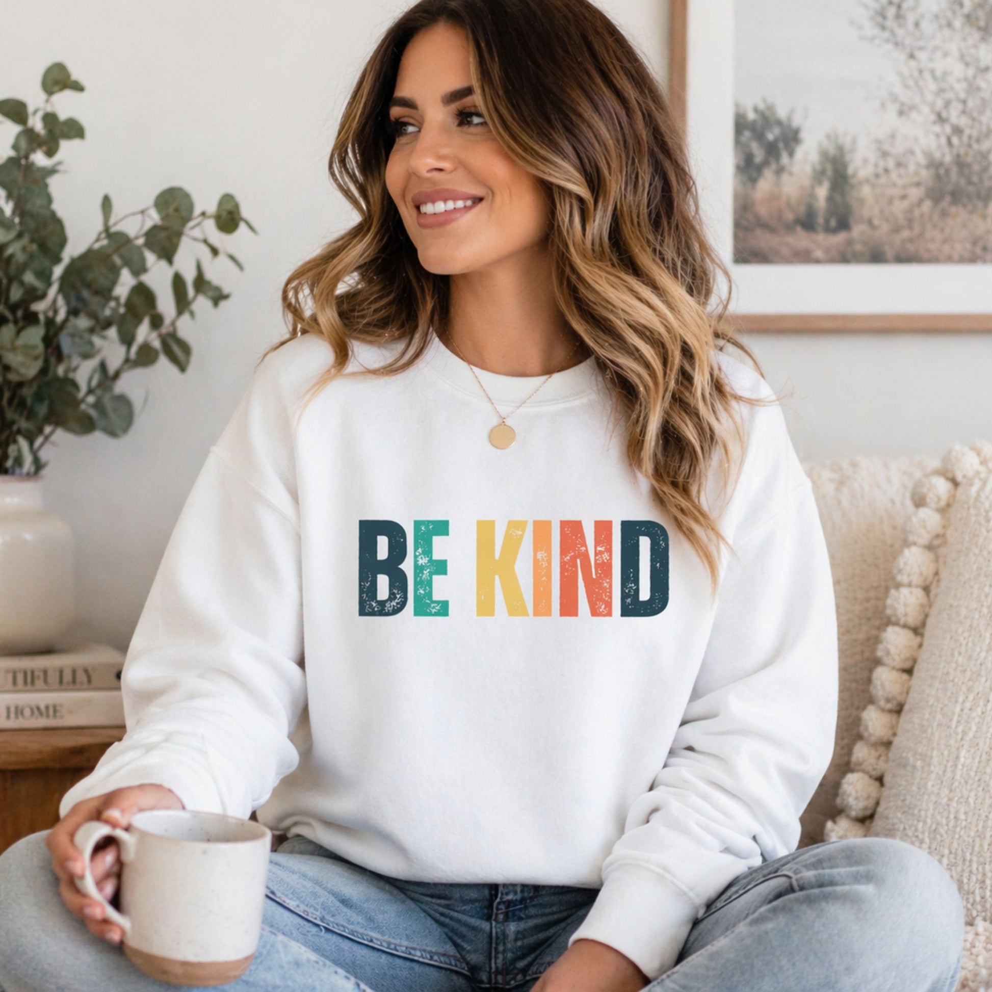 Woman sitting on a sofa with a cup of coffee wearing a white sweatshirt that says Be Kind in colorful distressed font.