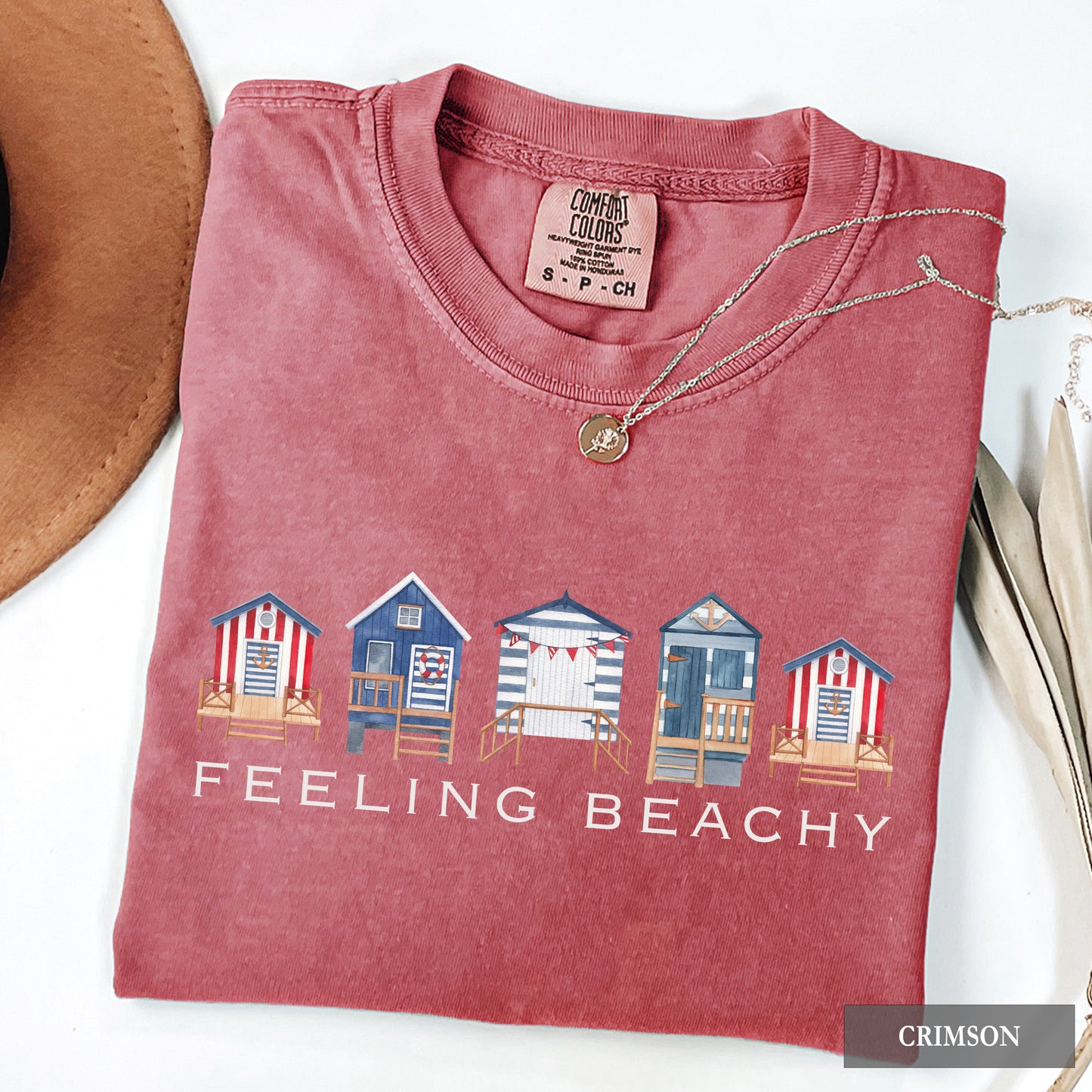 Folded red t-shirt with beach hut design and 'Feeling Beachy' text, next to a hat and necklace on a white background.