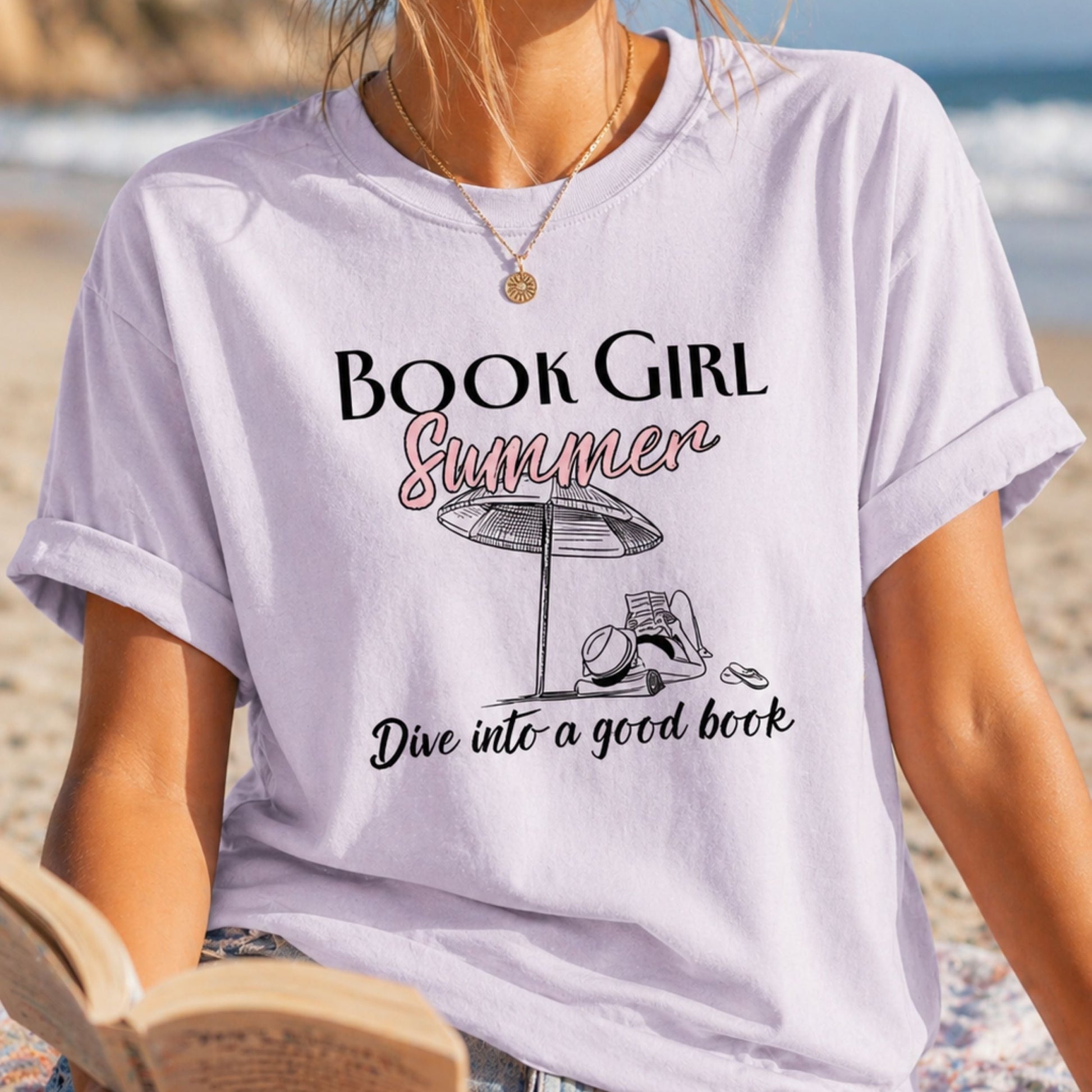 Woman on a beach wearing a 'Book Girl Summer' t-shirt and reading a book.