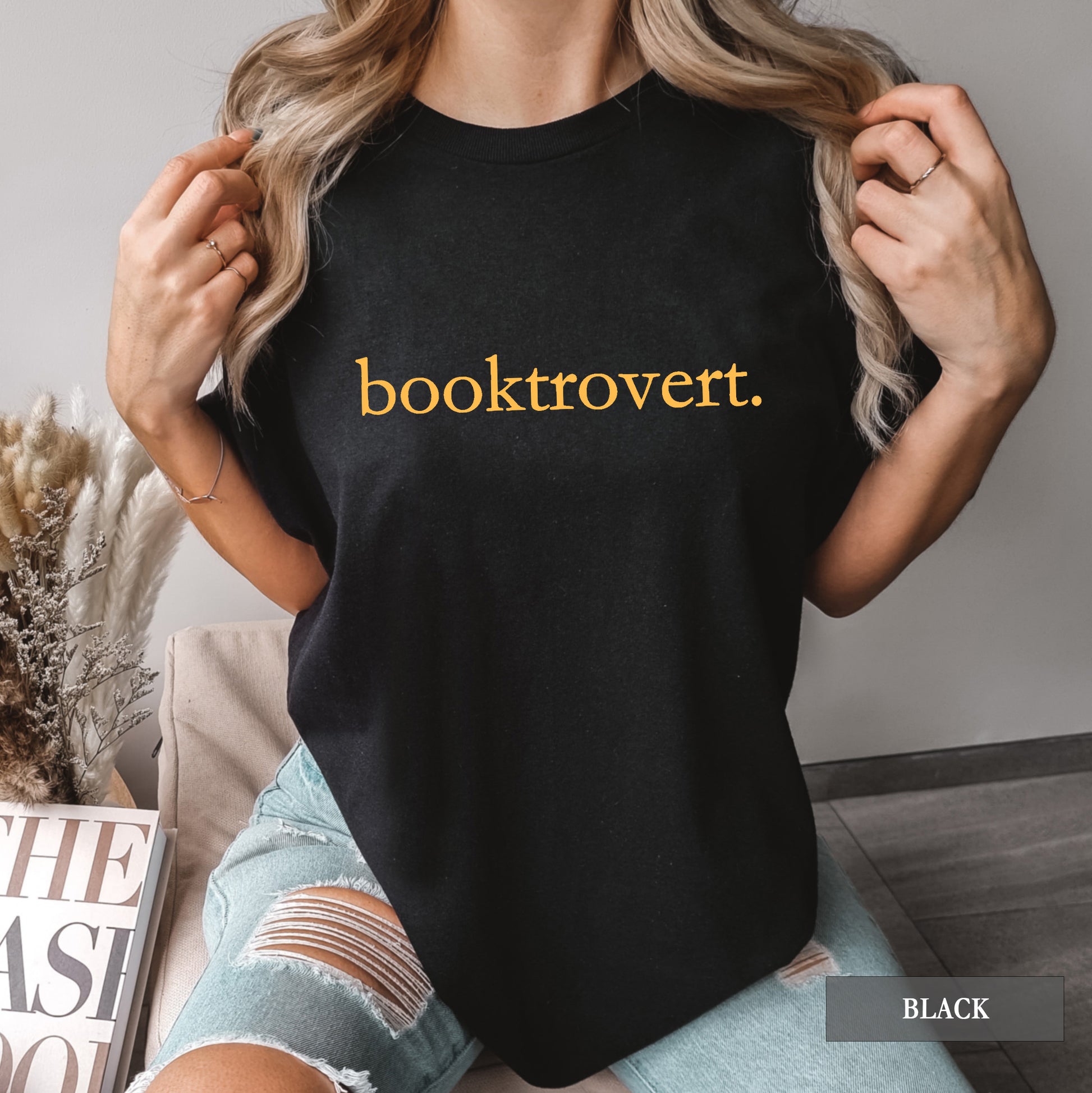 Woman wearing a black comfort colors shirt with "booktrovert" on the front in minimalist typewriter style font.