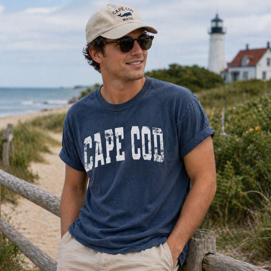 Man wearing a 'Cape Cod' t-shirt by the beach with a lighthouse in the background.